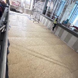 Usługi wykończeniowe - Montaż płyt OSB na podłodze w dużym pomieszczeniu. Widoczne rusztowania, instalacje pod sufitem i pracownik na drabinie. Etap prac wykończeniowych.