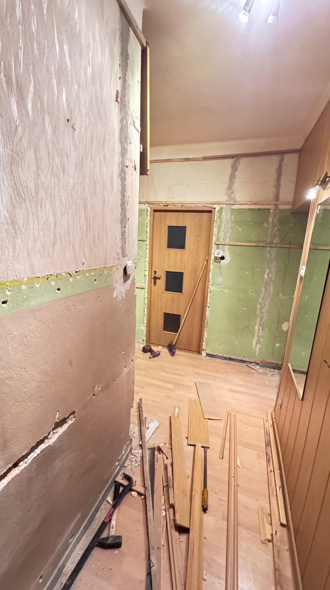 Korytarz w trakcie demontażu: zerwane tapety, odsłonięte ściany, widoczne narzędzia i fragmenty paneli podłogowych. Przygotowanie do remontu.