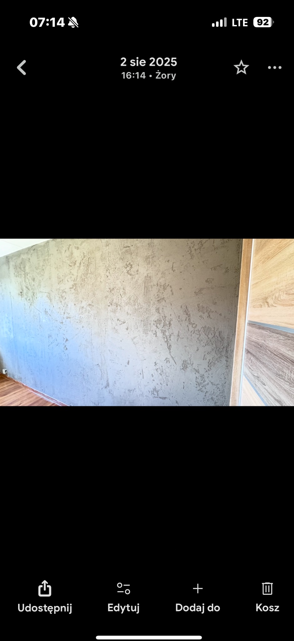 Ściana po szpachlowaniu z efektem dekoracyjnym, widoczna faktura i nierówności. Przy ścianie fragment drewnianych drzwi. Podłoga w ciepłym odcieniu brązu.