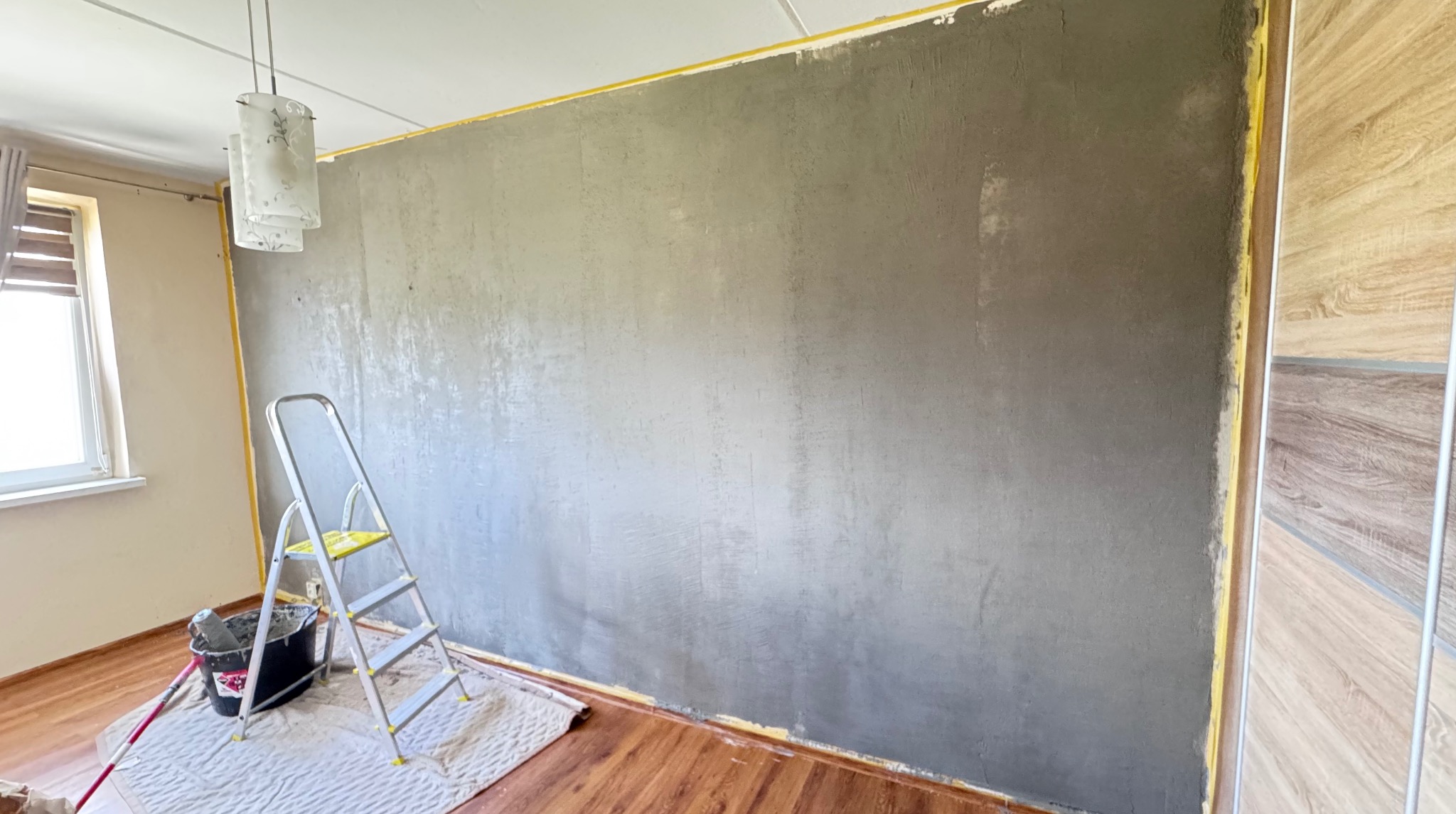 Ściana po szpachlowaniu, zabezpieczona taśmą malarską, z drabiną i akcesoriami malarskimi. Widoczna faktura szpachli i przygotowanie do dalszych prac wykończeniowych.