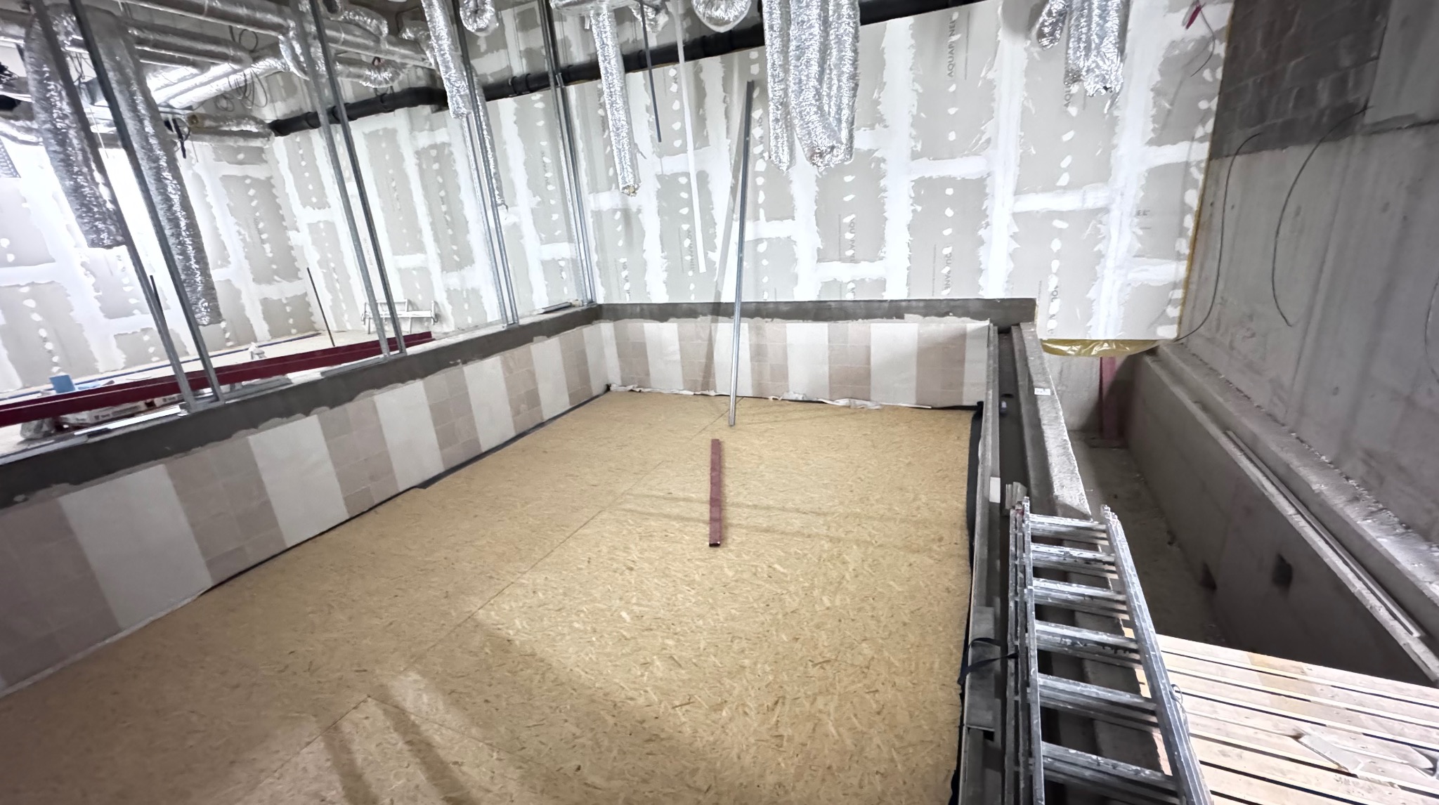 Wnętrze w trakcie wykańczania: ściany z płyt gipsowych po szpachlowaniu, częściowo wyłożona podłoga płytami OSB, widoczne instalacje wentylacyjne pod sufitem.