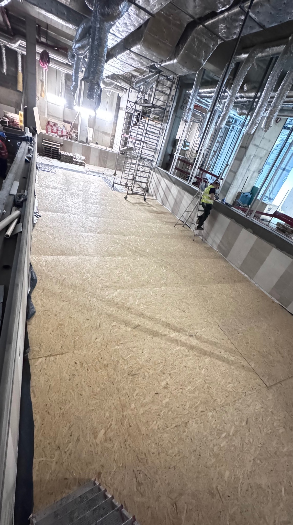 Montaż płyt OSB na podłodze w dużym pomieszczeniu. Widoczne rusztowania, instalacje pod sufitem i pracownik na drabinie. Etap prac wykończeniowych.