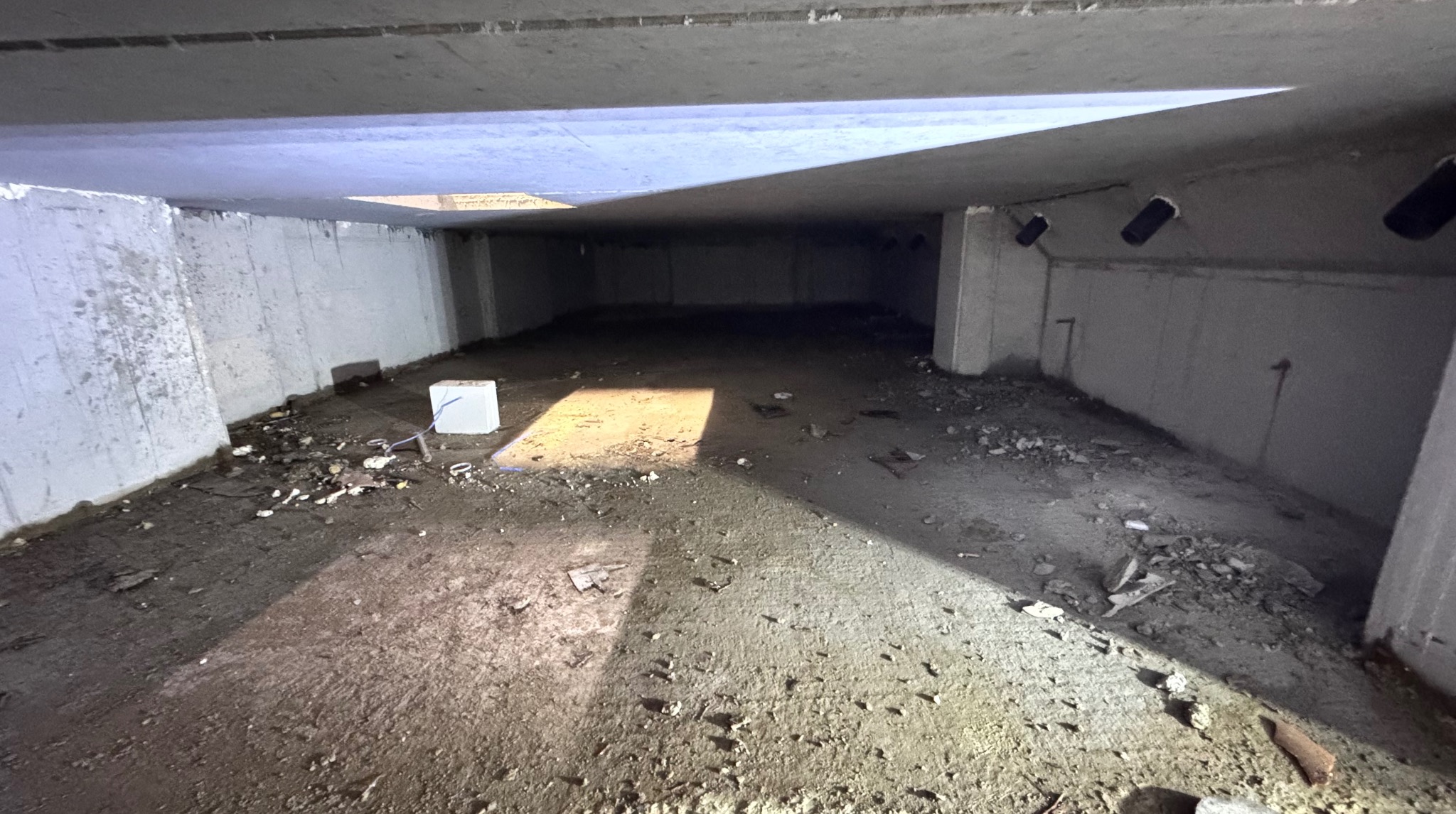 Surowe wnętrze piwnicy z betonowymi ścianami i podłogą, częściowo oświetlone naturalnym światłem wpadającym przez otwór w suficie. Widoczne ślady gruzu i pozostałości po pracach.