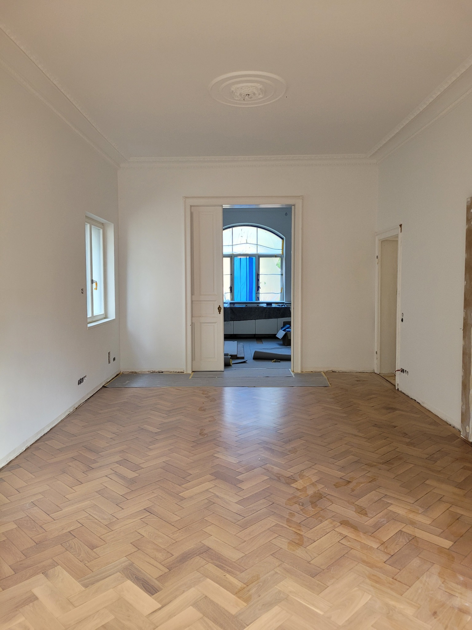 Jasne, przestronne wnętrze z parkietem ułożonym w jodełkę. Świeżo pomalowane ściany, widoczne listwy przypodłogowe w trakcie montażu. W tle widok na sąsiednie pomieszczenie.