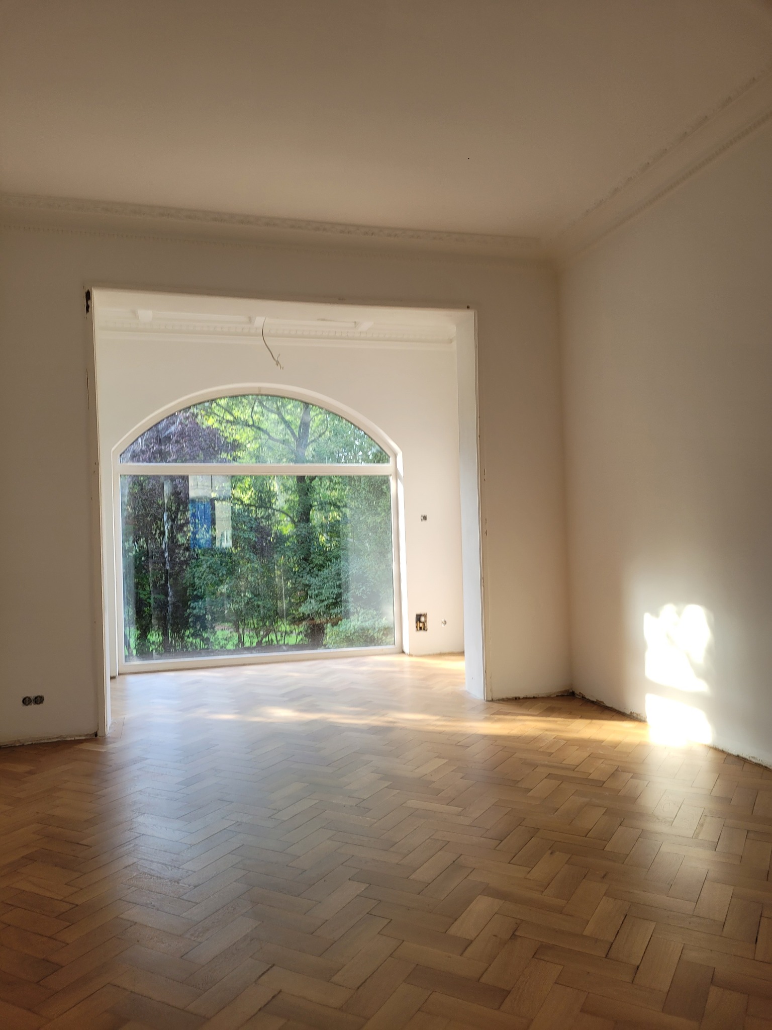 Jasne wnętrze z parkietem w jodełkę i dużym, łukowym oknem z widokiem na zieleń. Ściany białe, gładkie, z dekoracyjnym gzymsem pod sufitem. Częściowo odnowione.