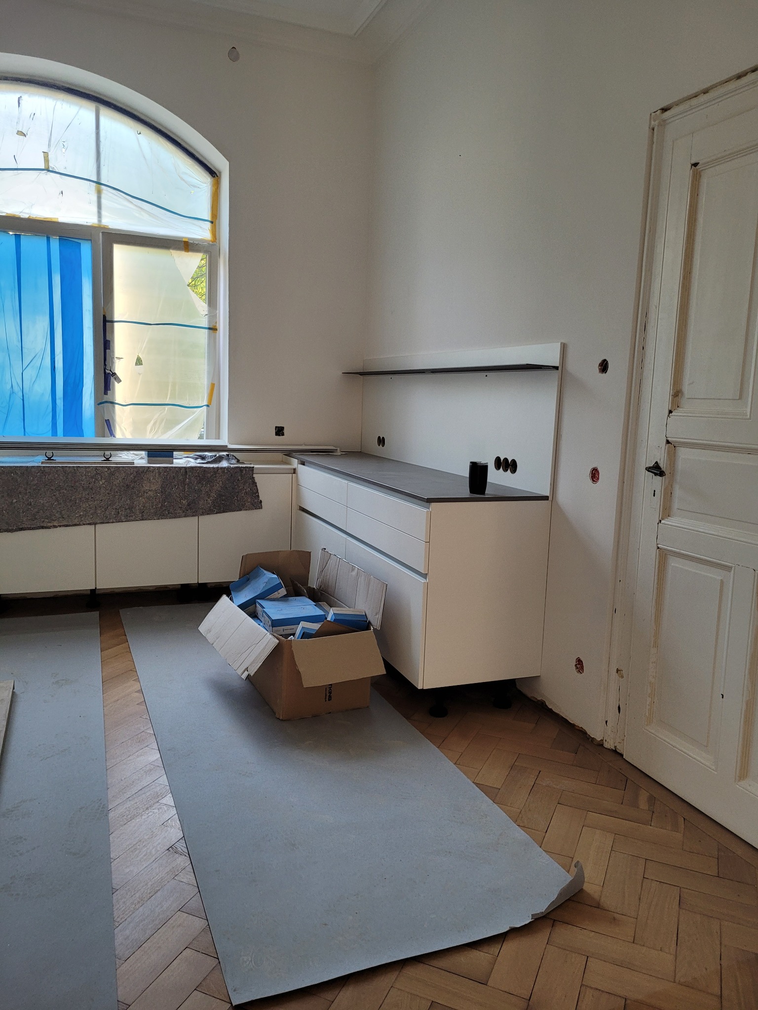 Montaż białej zabudowy kuchennej z szarym blatem w trakcie wykańczania. Okno zasłonięte folią. Podłoga częściowo zabezpieczona szarą płytą. Widoczna skrzynka z narzędziami.