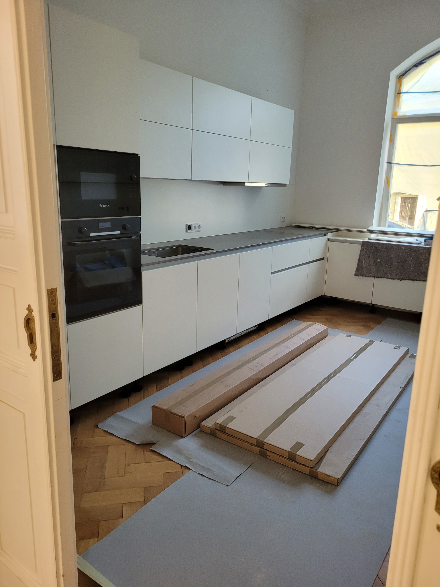 Jasna kuchnia w trakcie montażu. Widoczne białe szafki, sprzęt AGD i elementy do zabudowy na podłodze. Parkiet w jodełkę, okno z osłoną. Ujęcie zza uchylonych drzwi.