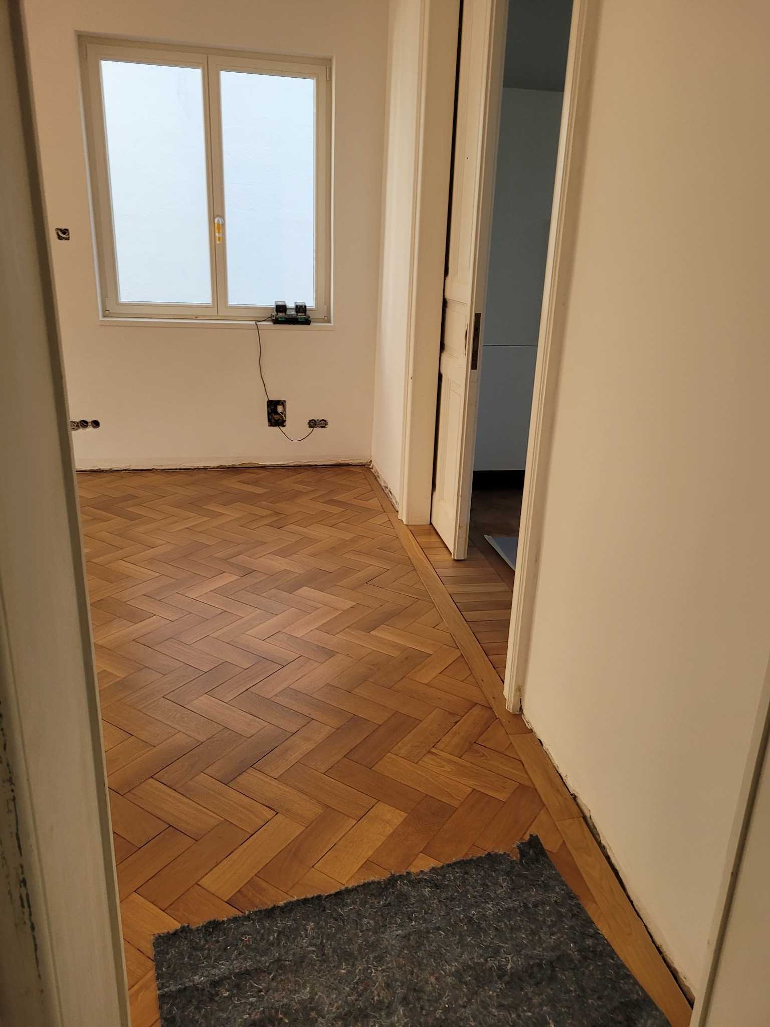 Ukończona podłoga z parkietu ułożonego w jodełkę, widok zza otwartych drzwi. Widoczne listwy przypodłogowe częściowo zdjęte, okno z białymi ramami.