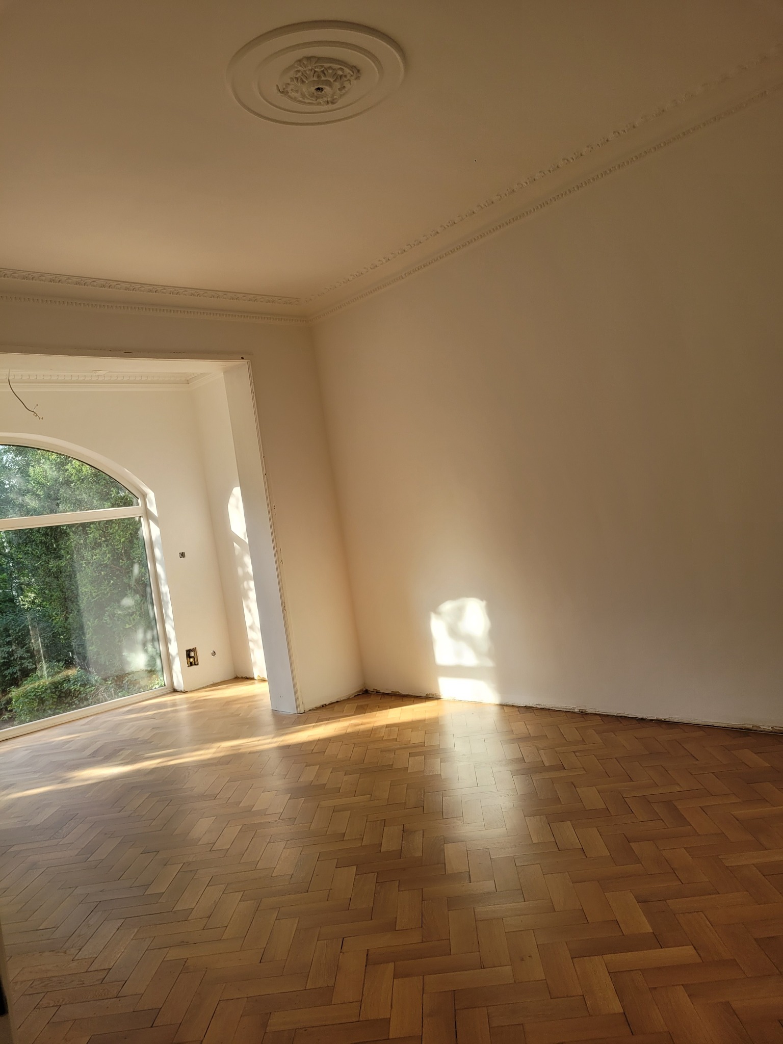 Pusty pokój z parkietem w jodełkę, listwami dekoracyjnymi pod sufitem i dużym oknem łukowym. Świeże, białe ściany, gotowe do dalszych prac wykończeniowych.