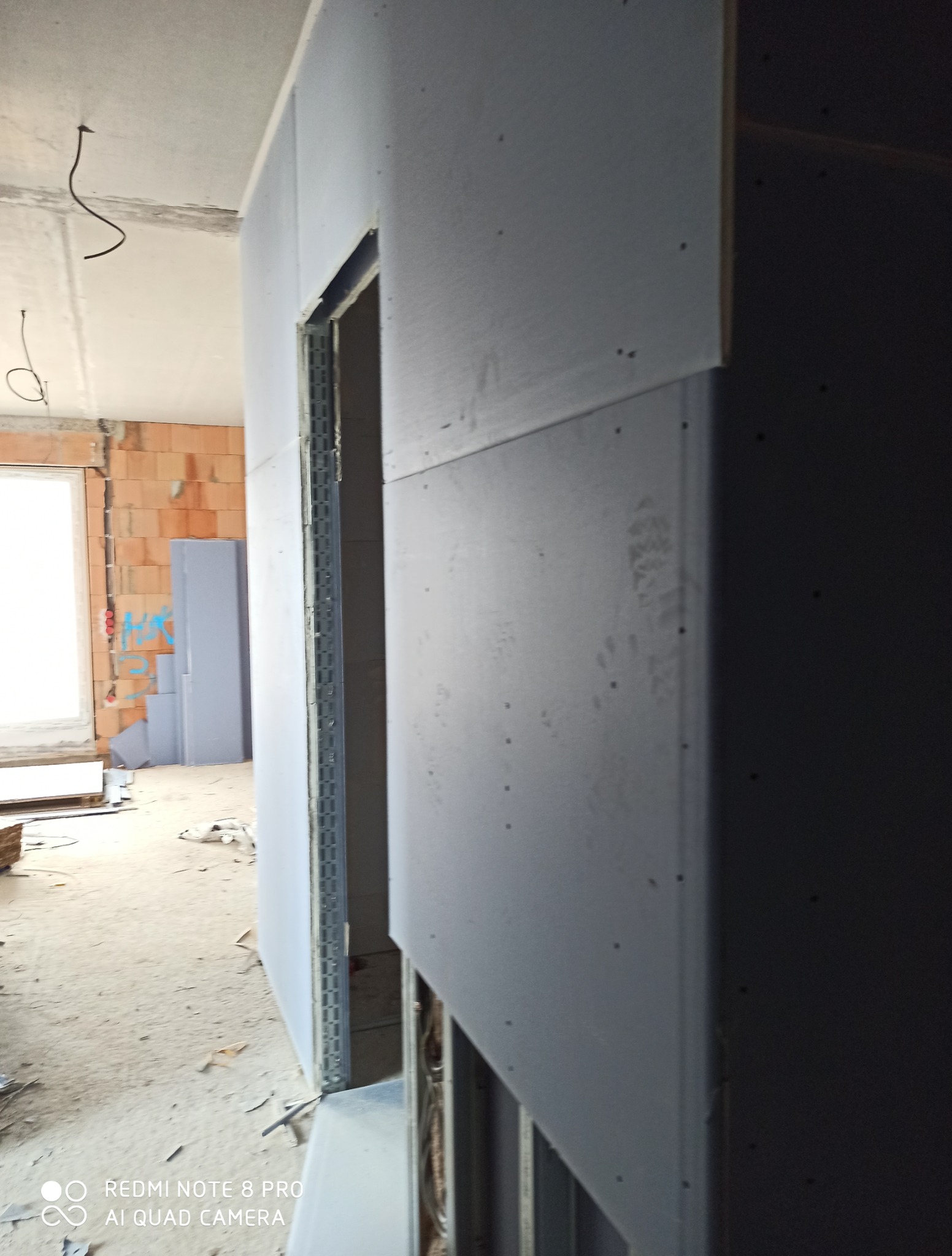 Montaż płyt karton-gips na metalowym stelażu w surowym wnętrzu. Widoczne przewody elektryczne i fragment okna. Ściany z czerwonej cegły w tle.