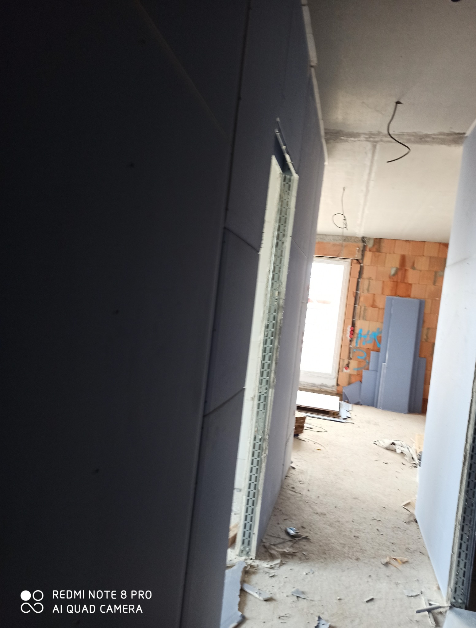 Montaż płyt karton-gips na ścianie z widocznym profilem metalowym przy otworze drzwiowym. W tle surowy mur z cegieł i kable. W pomieszczeniu widoczny bałagan.