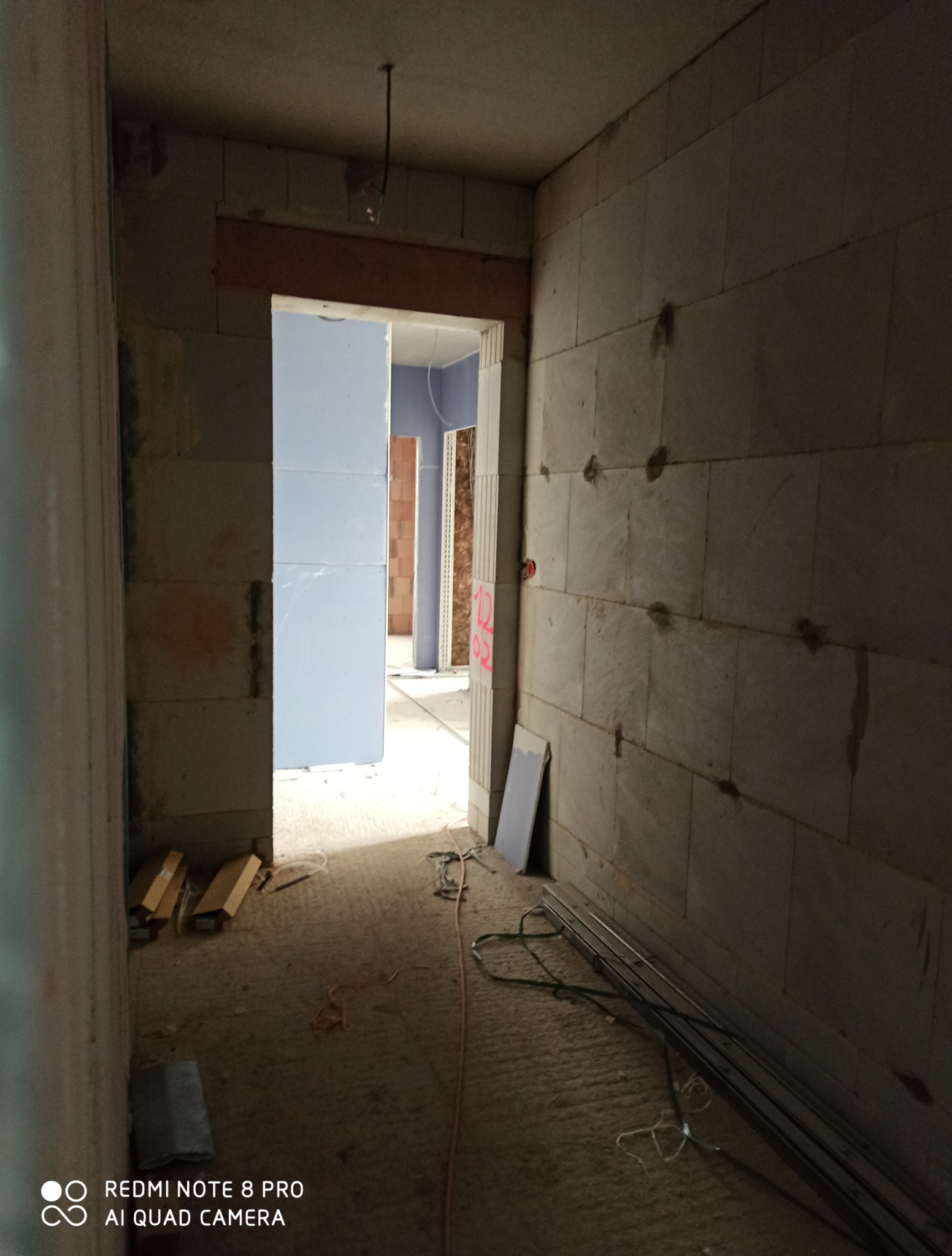 Wnętrze w trakcie remontu: surowe ściany z bloczków, widoczne instalacje, przygotowane otwory drzwiowe, płyty gipsowe. W tle kolejne pomieszczenia w stanie surowym.