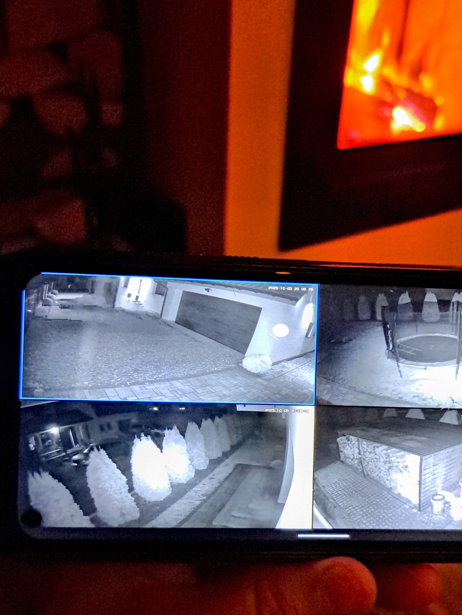 Podgląd z kamer monitoringu na smartfonie: brama garażowa, podwórko, rząd drzew i drewutnia, wszystko w czerni i bieli, datowane. W tle kominek.