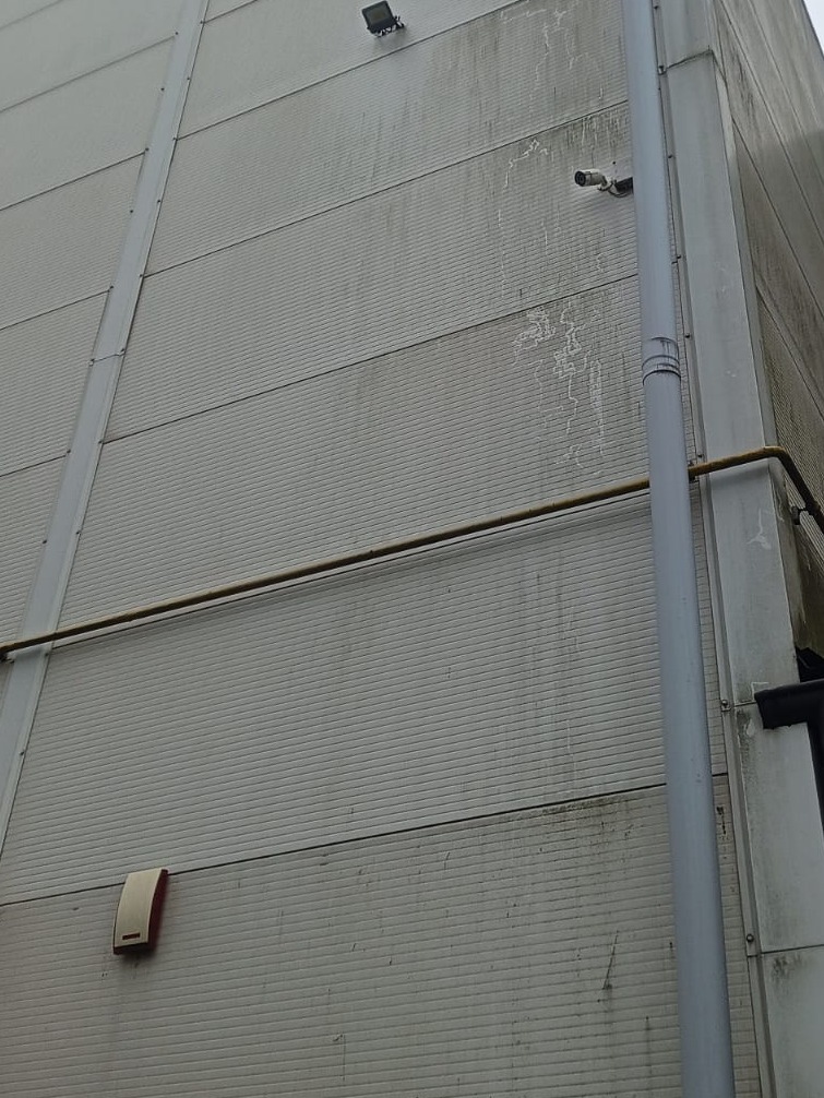 Elewacja budynku z zamontowaną kamerą przemysłową, lampą oświetleniową i rurą spustową. Widoczne zabrudzenia na panelach ściennych.