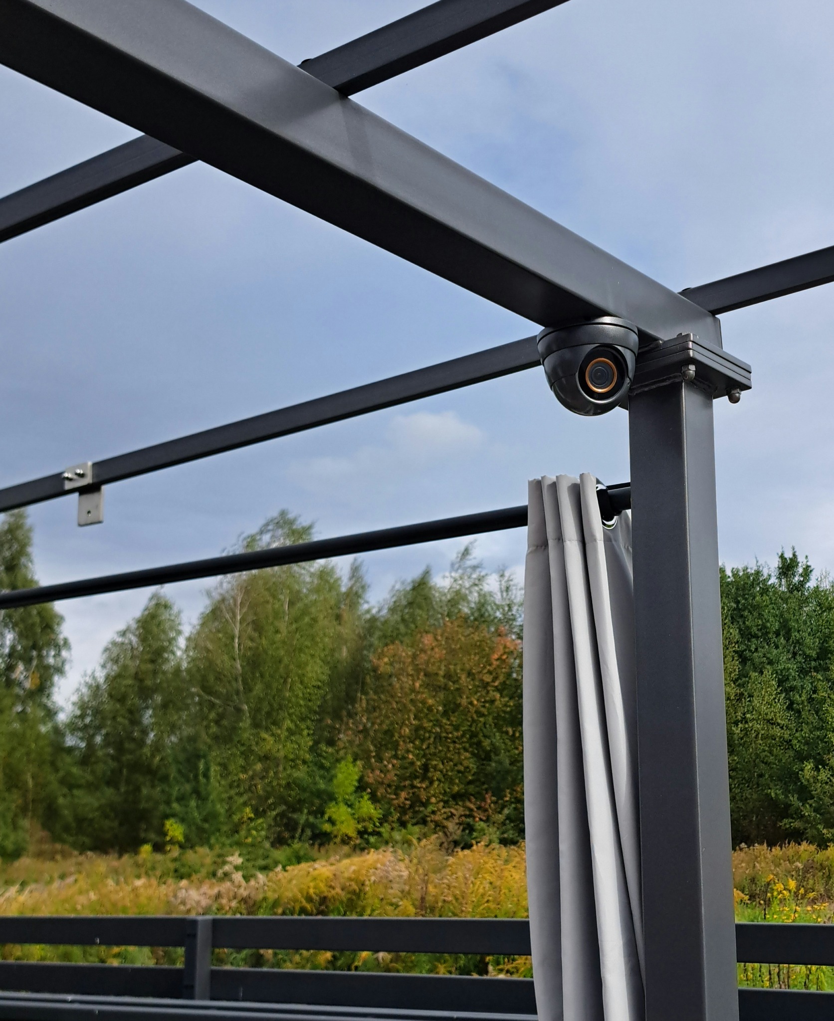 Kamera kopułkowa monitoringu zamontowana na konstrukcji z zasłonami w tle zieleni. Widok na nowoczesny system monitoringu zewnętrznego.