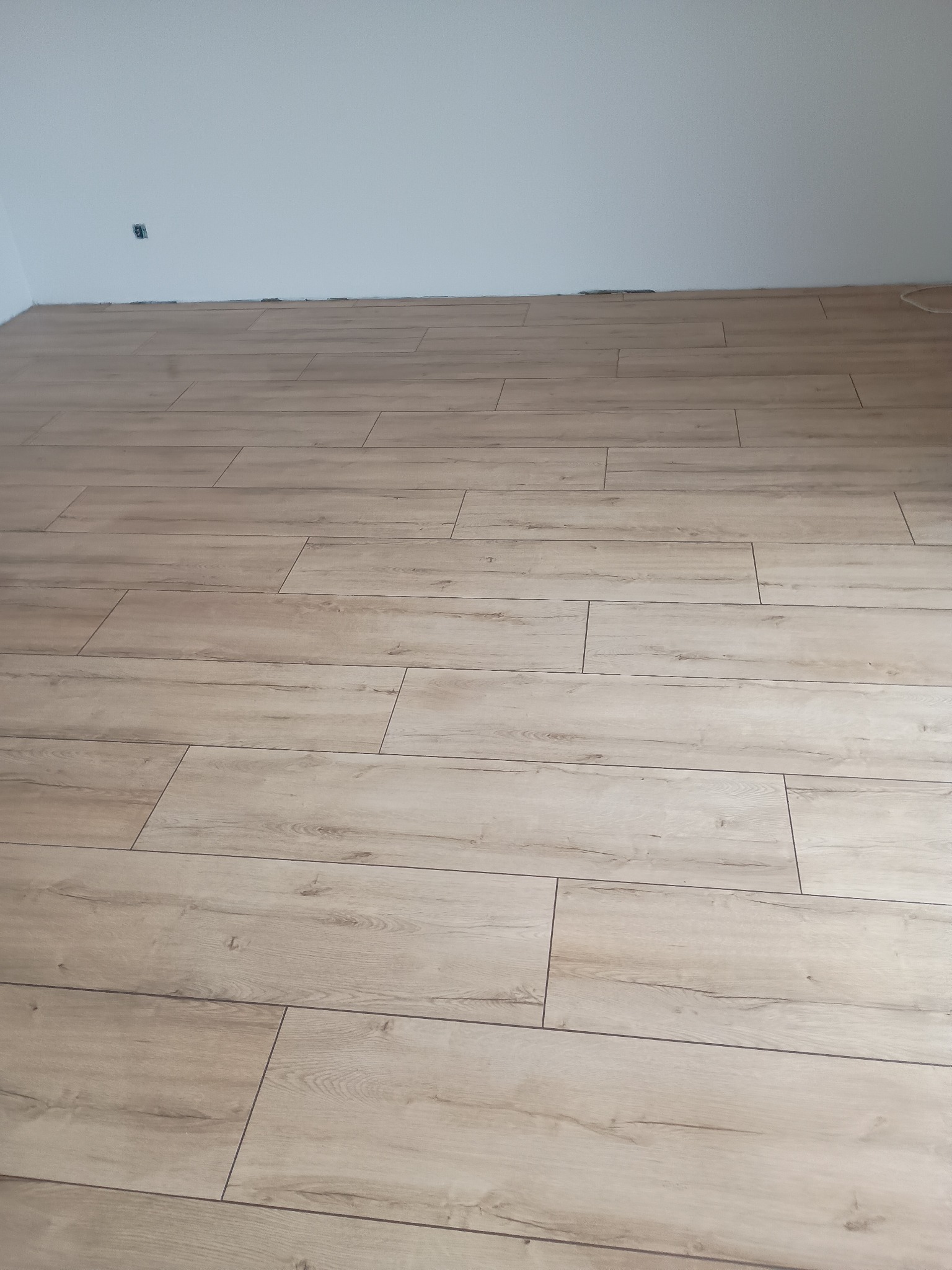 Ułożona podłoga z paneli imitujących drewno w jasnym odcieniu, widok z góry, fragment ściany z gniazdkiem w tle. Nowe wykończenie podłogi.