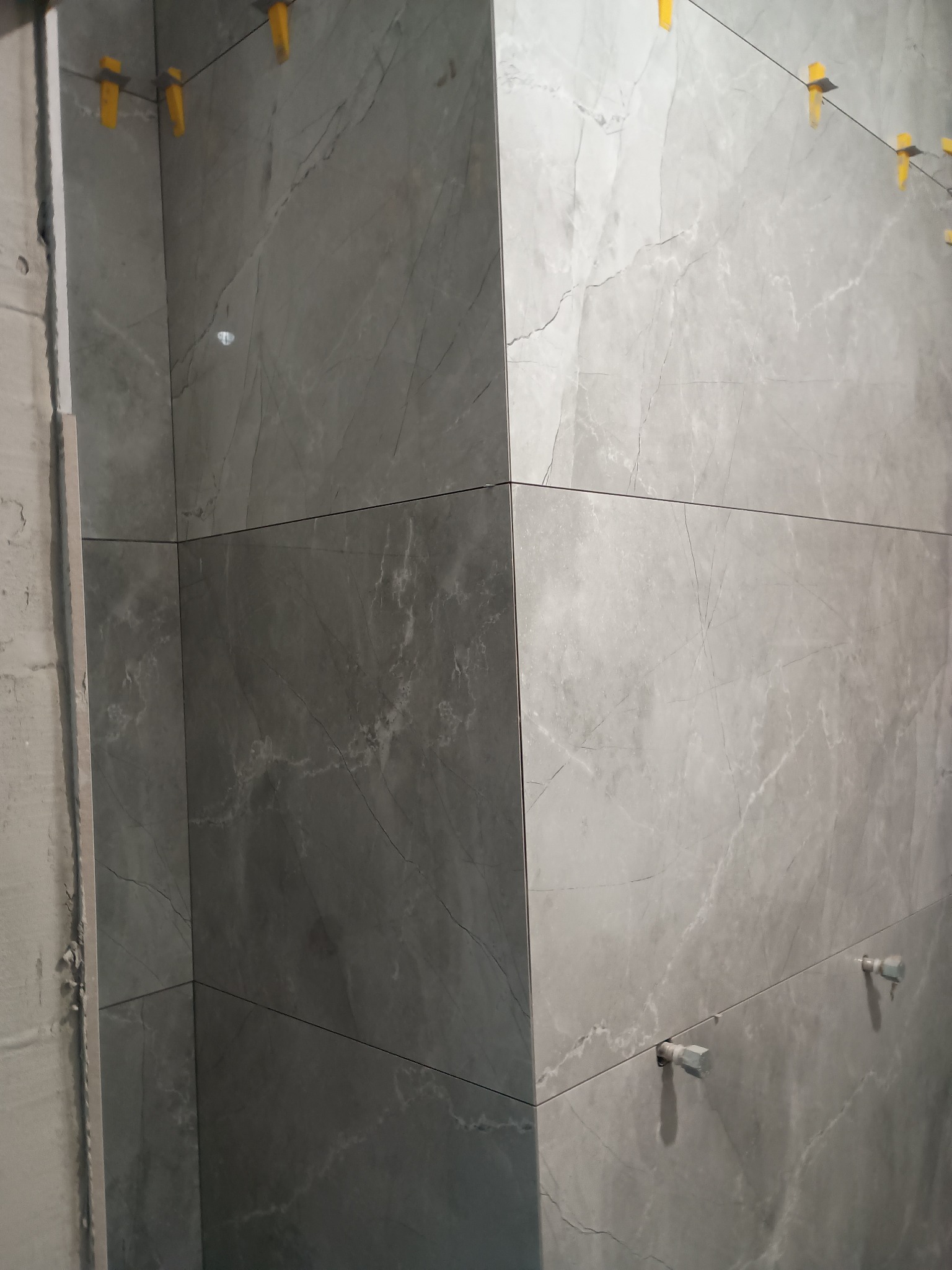 Szare płytki imitujące marmur na ścianie w trakcie montażu z użyciem żółtych krzyżyków dystansowych i białych klinów poziomujących.
