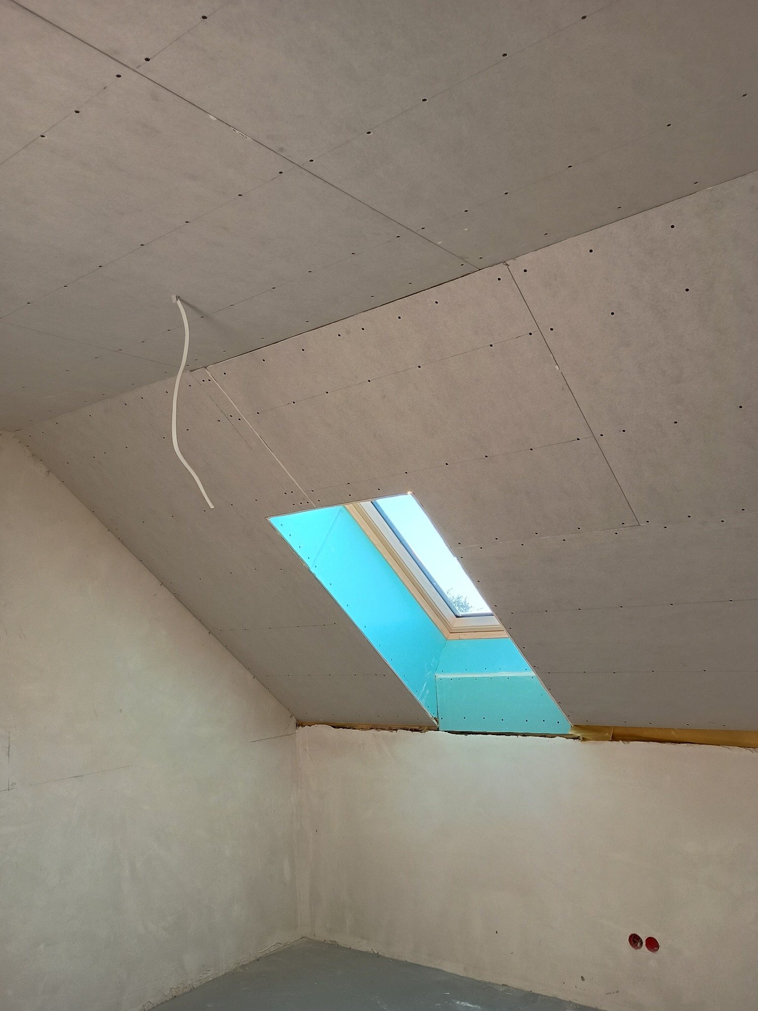 Poddasze w trakcie wykańczania: zamontowane płyty karton-gips na skosach, widoczne okno dachowe i wystający kabel. Ściany częściowo wyszpachlowane.