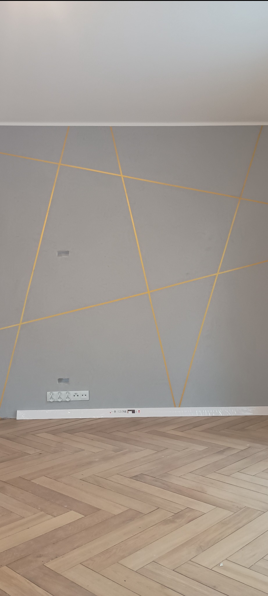 Szara ściana z geometrycznym wzorem z złotych linii, gniazdka elektryczne i fragment podłogi z parkietem w jodełkę. Listwa przypodłogowa w folii leży na podłodze.