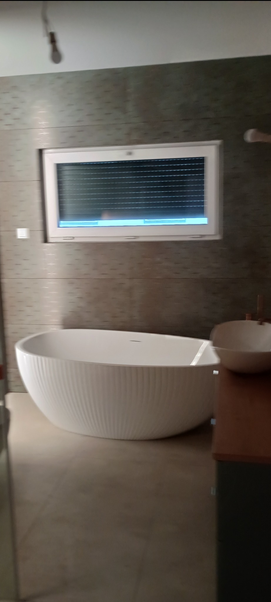 Nowoczesna łazienka z wolnostojącą wanną o żłobkowanej powierzchni, umywalką na blacie i oknem z żaluzjami. Stonowana kolorystyka płytek.