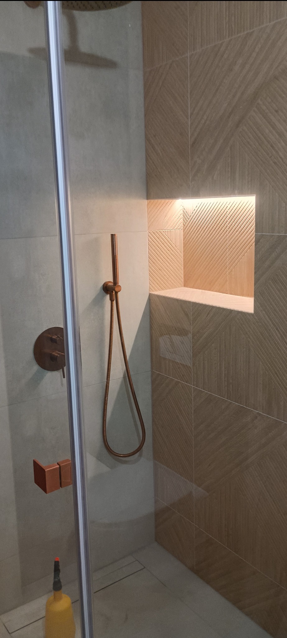 Nowoczesna kabina prysznicowa z miedzianą armaturą i wnęką w ścianie wyłożoną płytkami z subtelnym wzorem. Minimalistyczny design, stonowana kolorystyka.