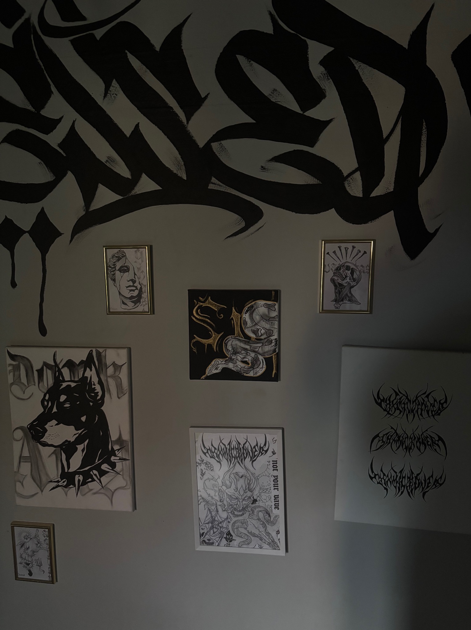 Ściana z różnorodnymi, czarno-białymi grafikami i rysunkami w ramach i bez, w tym graffiti na górze i szkice zwierząt/postaci na dole.