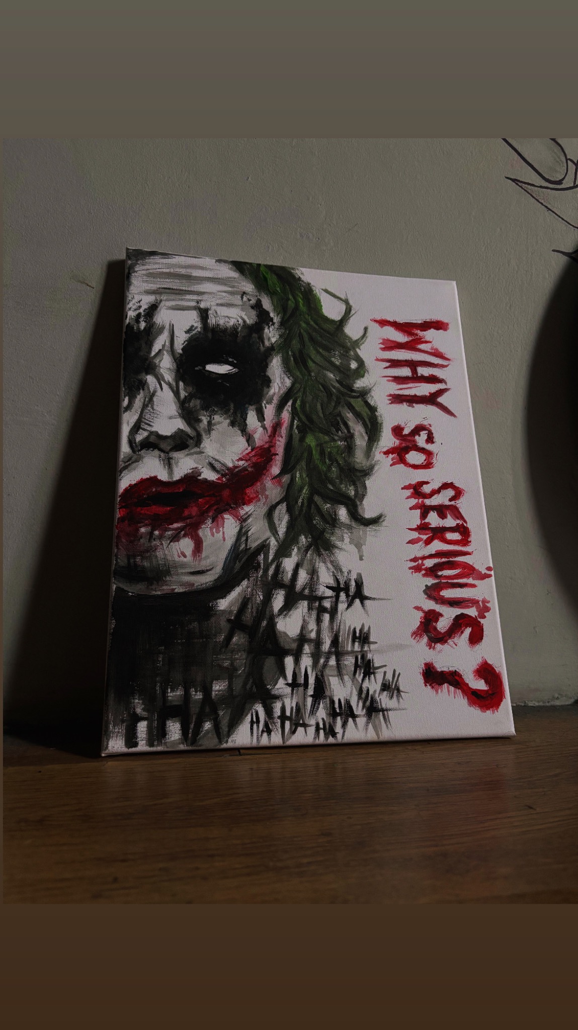 Ręcznie malowany portret Jokera na płótnie, z napisem 'Why so serious?' w czerwieni, stoi na drewnianym blacie na tle szarej ściany, artystyczny styl.