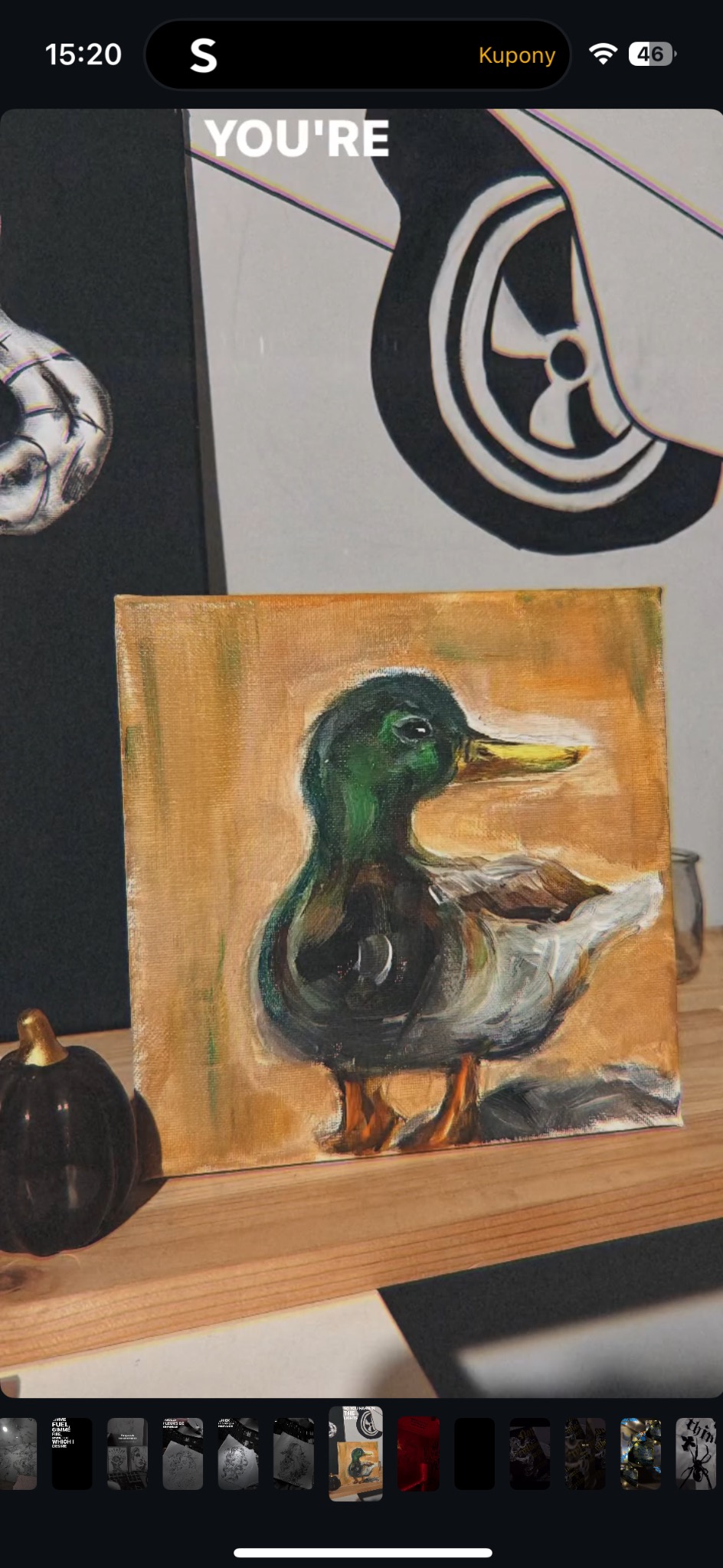 Ręcznie malowany obraz olejny kaczki na płótnie, ustawiony na drewnianej półce obok małej, czarnej dyni. W tle inne obrazy.