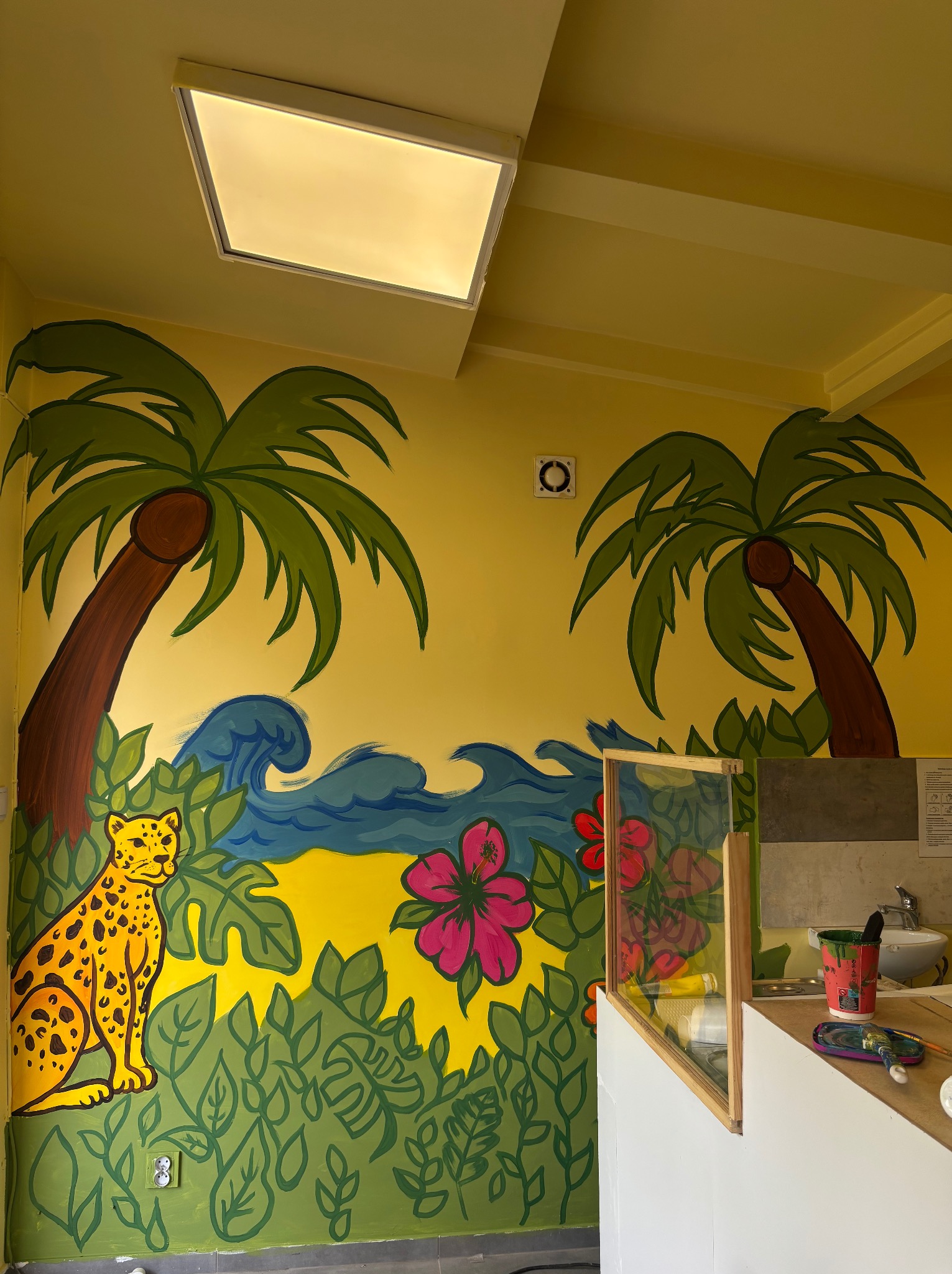 Żółta ściana z ręcznie malowanym tropikalnym motywem: palmy, fale, kwiaty hibiskusa i siedzący gepard. Widoczne narzędzia malarskie.