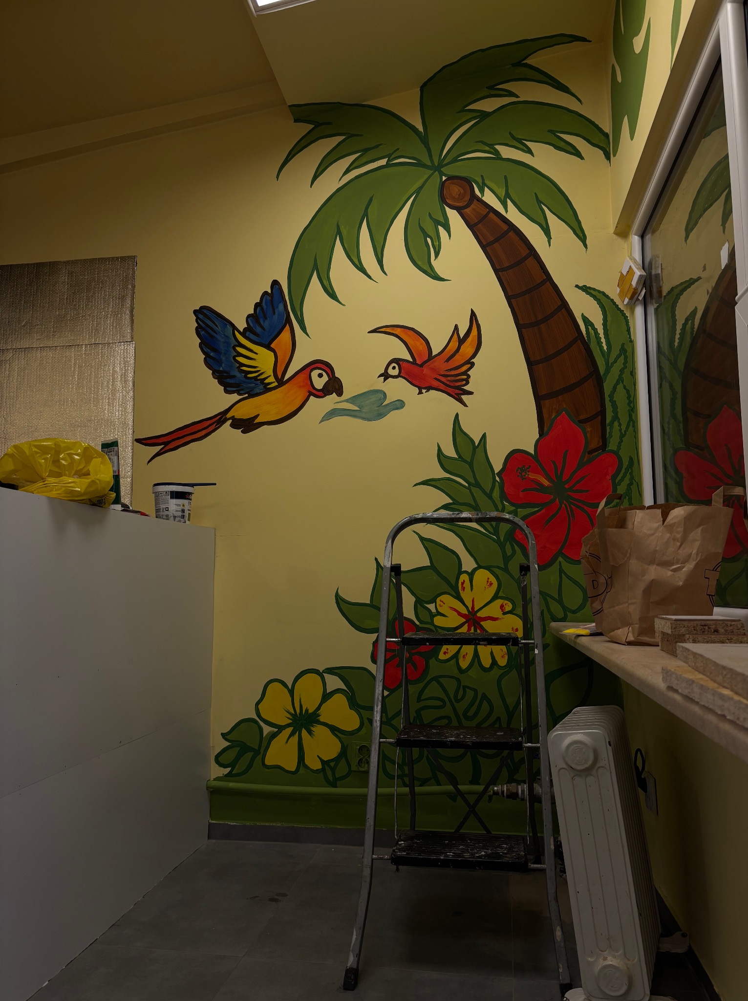 Kolorowa, ręcznie malowana ściana z motywem tropikalnym: palma, kwiaty i papugi. W tle drabina i akcesoria malarskie, co sugeruje etap pracy nad malowidłem.