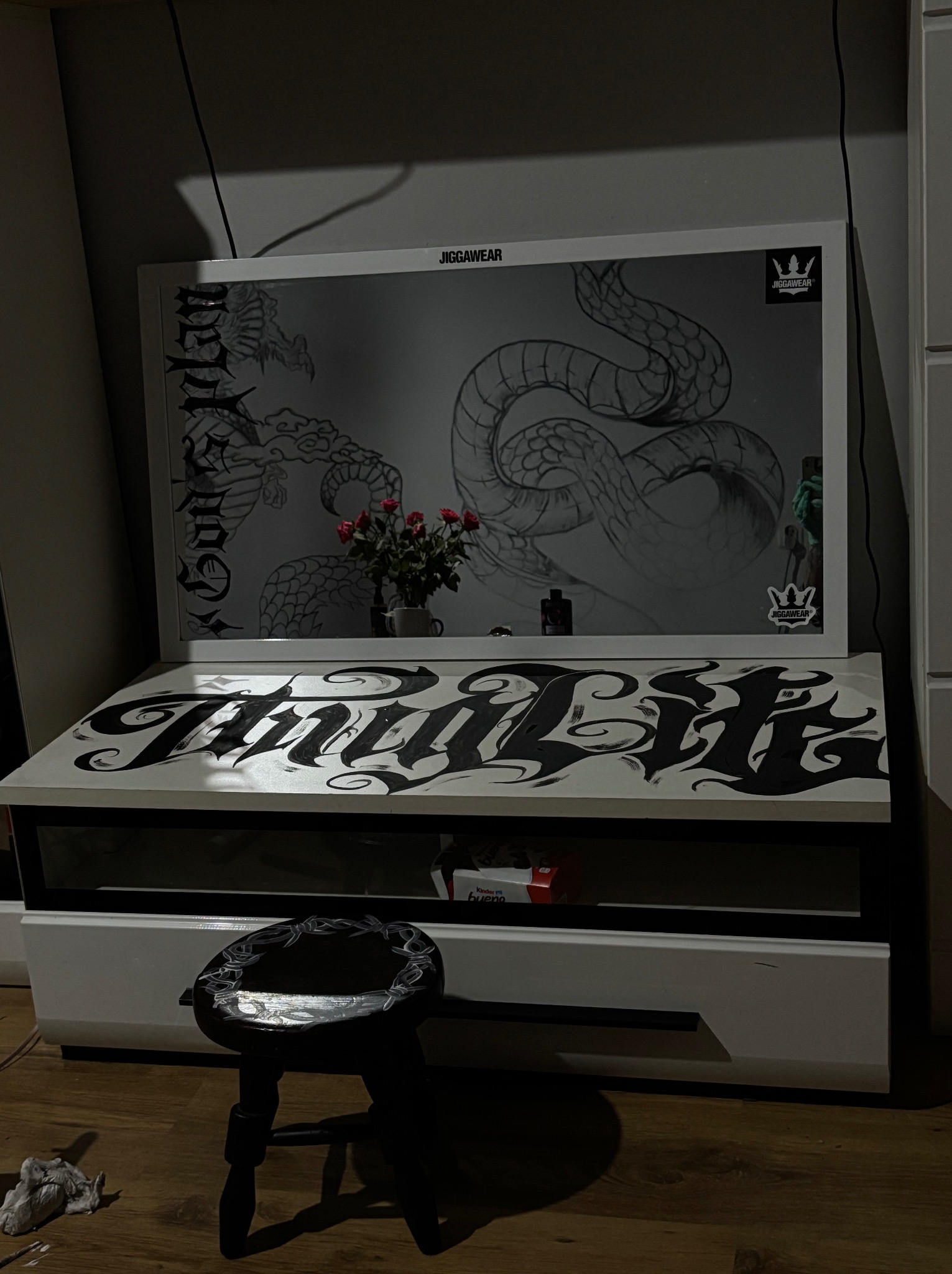Stanowisko pracy artysty z rysunkami węży, logotypami JiggaWear i napisem 'Final Life' na biurku. Mały stołek obok. Całość w stonowanej kolorystyce i ciemnym oświetleniu.