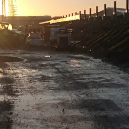 Teren budowy o zmierzchu: błotnista droga, sprzęt budowlany w tle, słupy wysokiego napięcia i zachodzące słońce. Specyficzne oświetlenie.