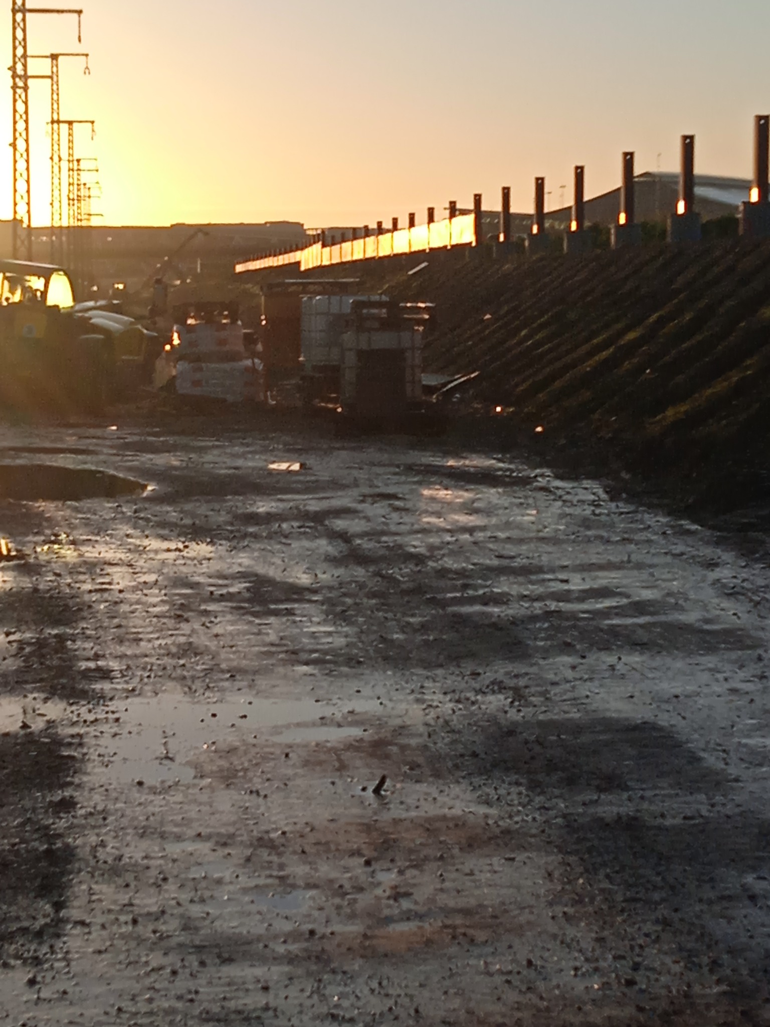 Teren budowy o zmierzchu: błotnista droga, sprzęt budowlany w tle, słupy wysokiego napięcia i zachodzące słońce. Specyficzne oświetlenie.