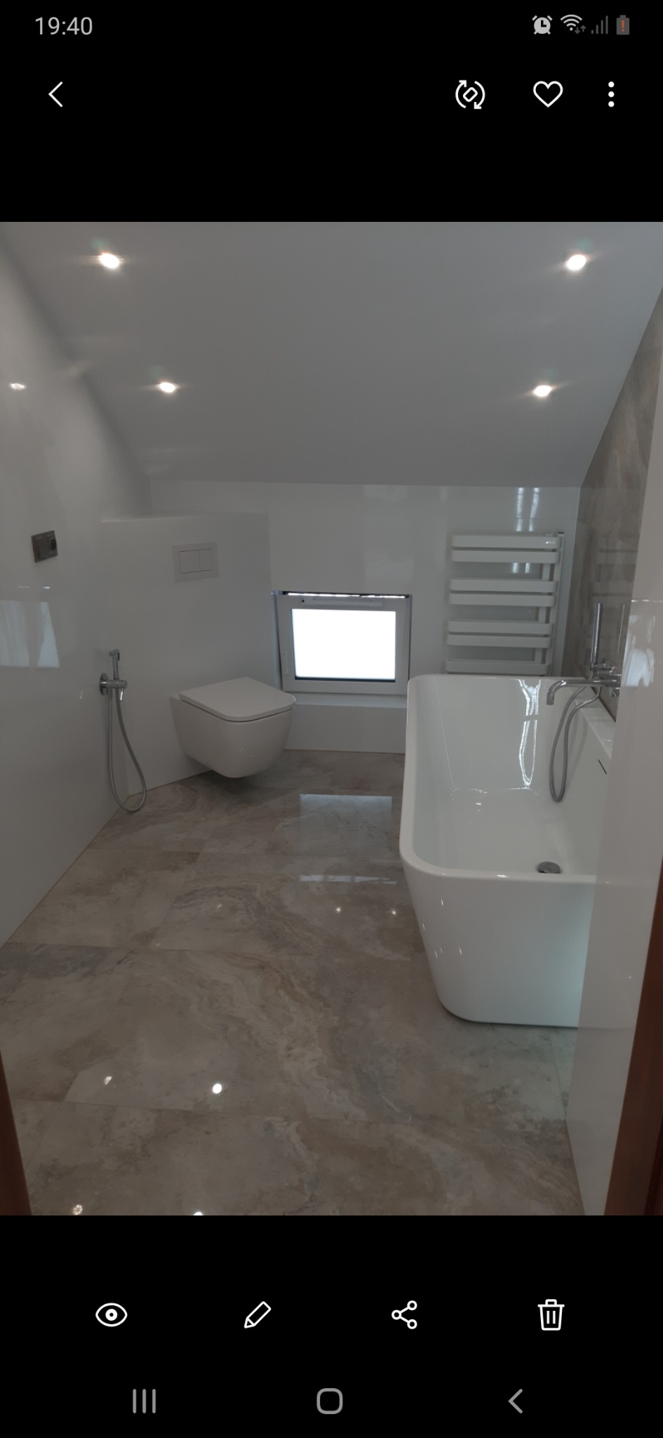 Nowoczesna łazienka z wolnostojącą wanną, podwieszaną toaletą i beżowymi płytkami podłogowymi. Minimalistyczny design z oknem w ścianie.