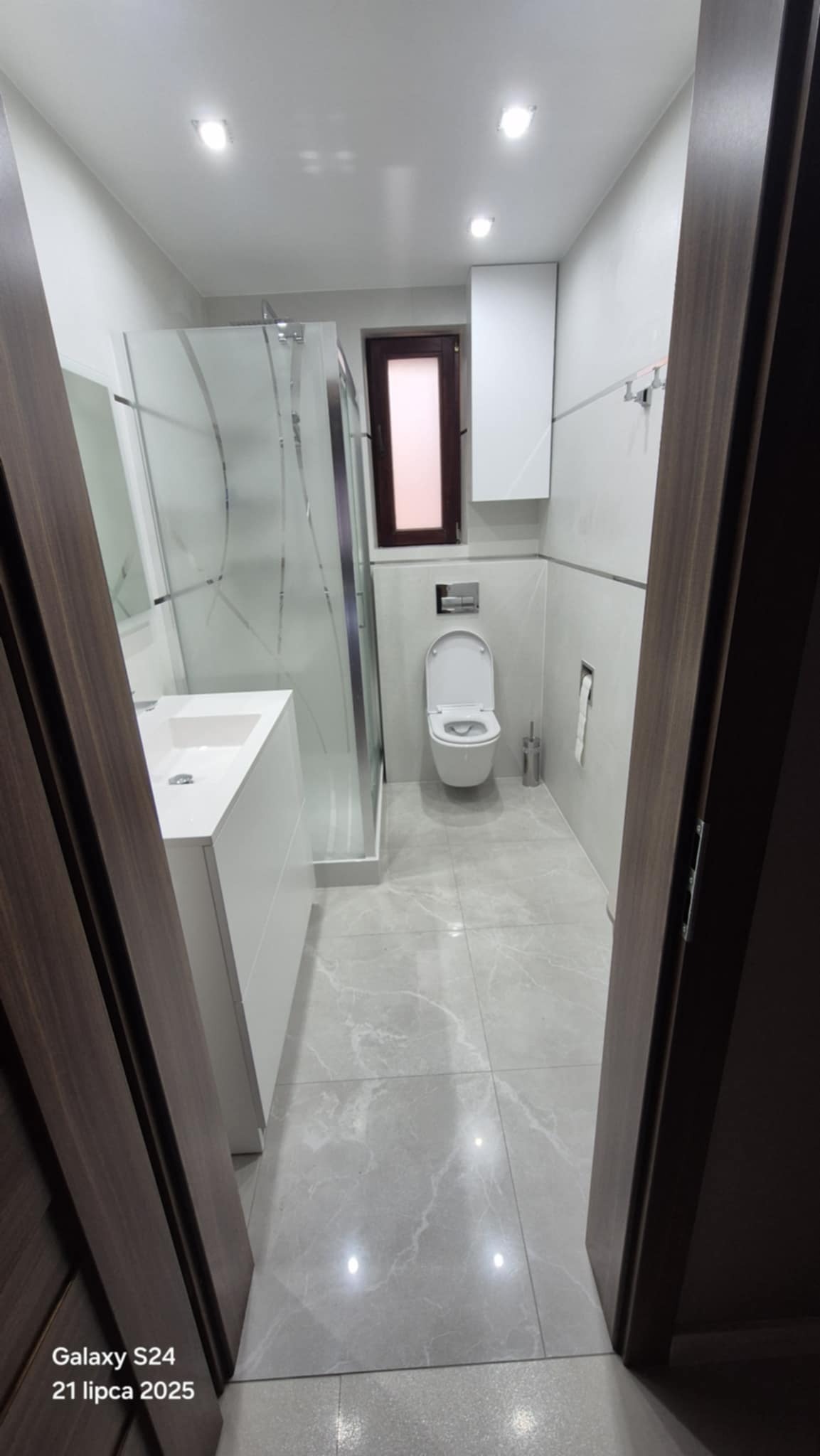 Nowoczesna łazienka z kabiną prysznicową, umywalką z szafką i toaletą. Jasne płytki podłogowe i ścienne. Widok zza otwartych, ciemnych drzwi.