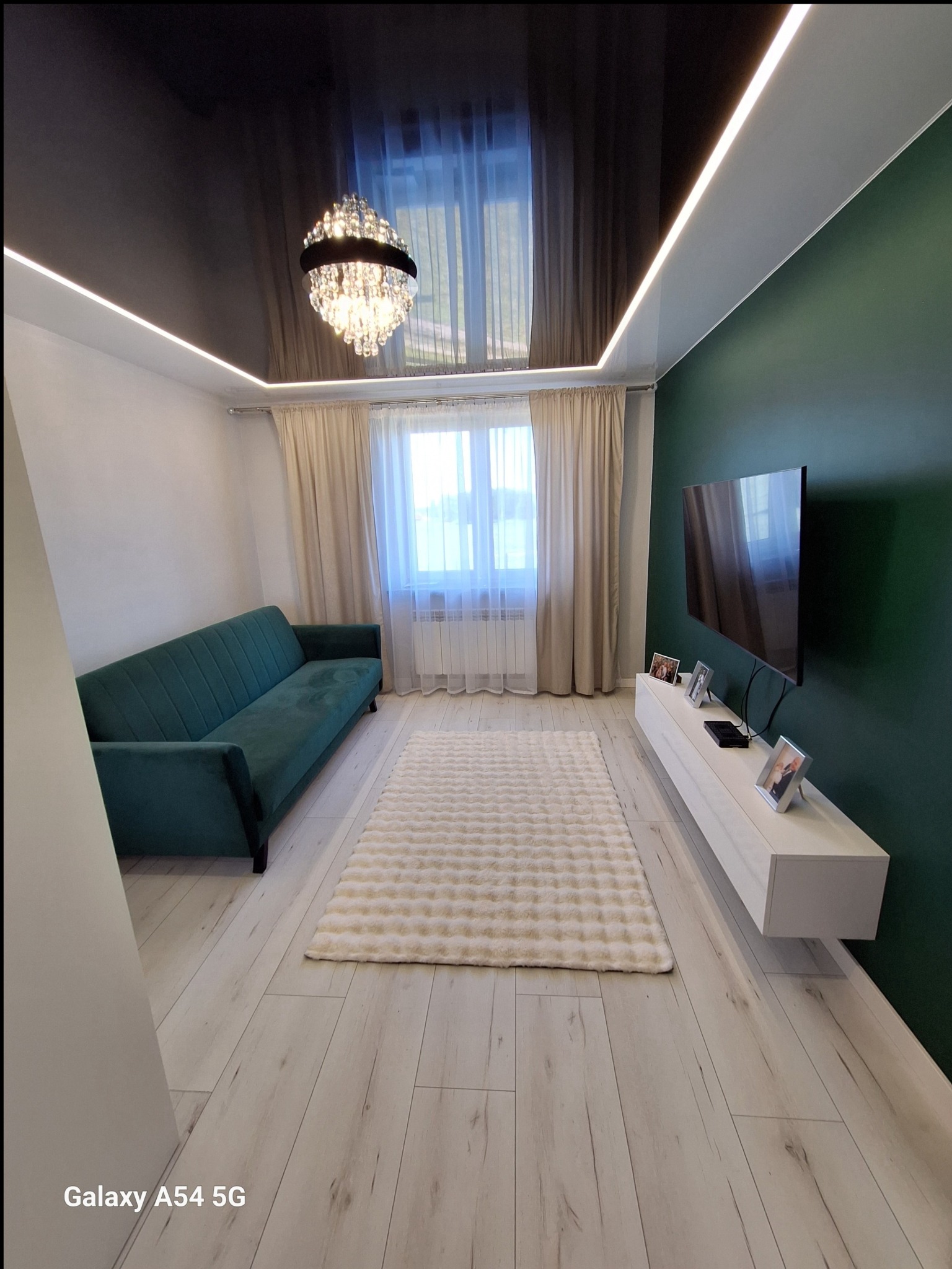 Salon po remoncie: zielona ściana, białe listwy LED, czarny sufit lustrzany, kryształowy żyrandol, turkusowa sofa i puszysty dywan na jasnej podłodze.