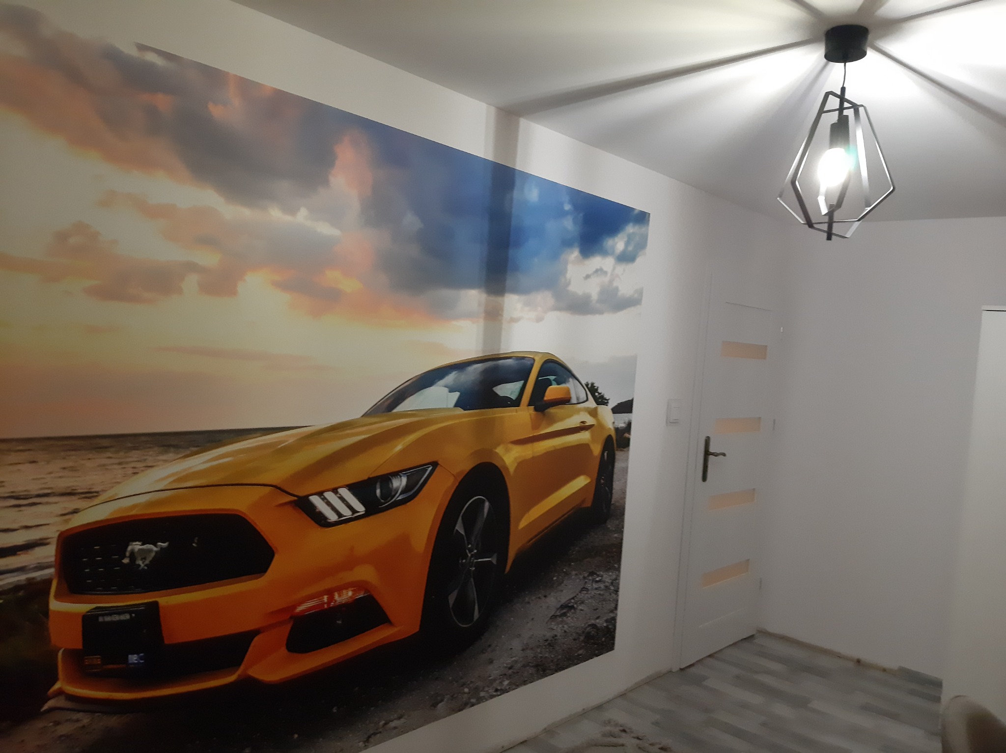 Fototapeta z motywem żółtego samochodu sportowego na tle zachodzącego słońca, umieszczona na ścianie w jasnym wnętrzu z nowoczesną lampą.