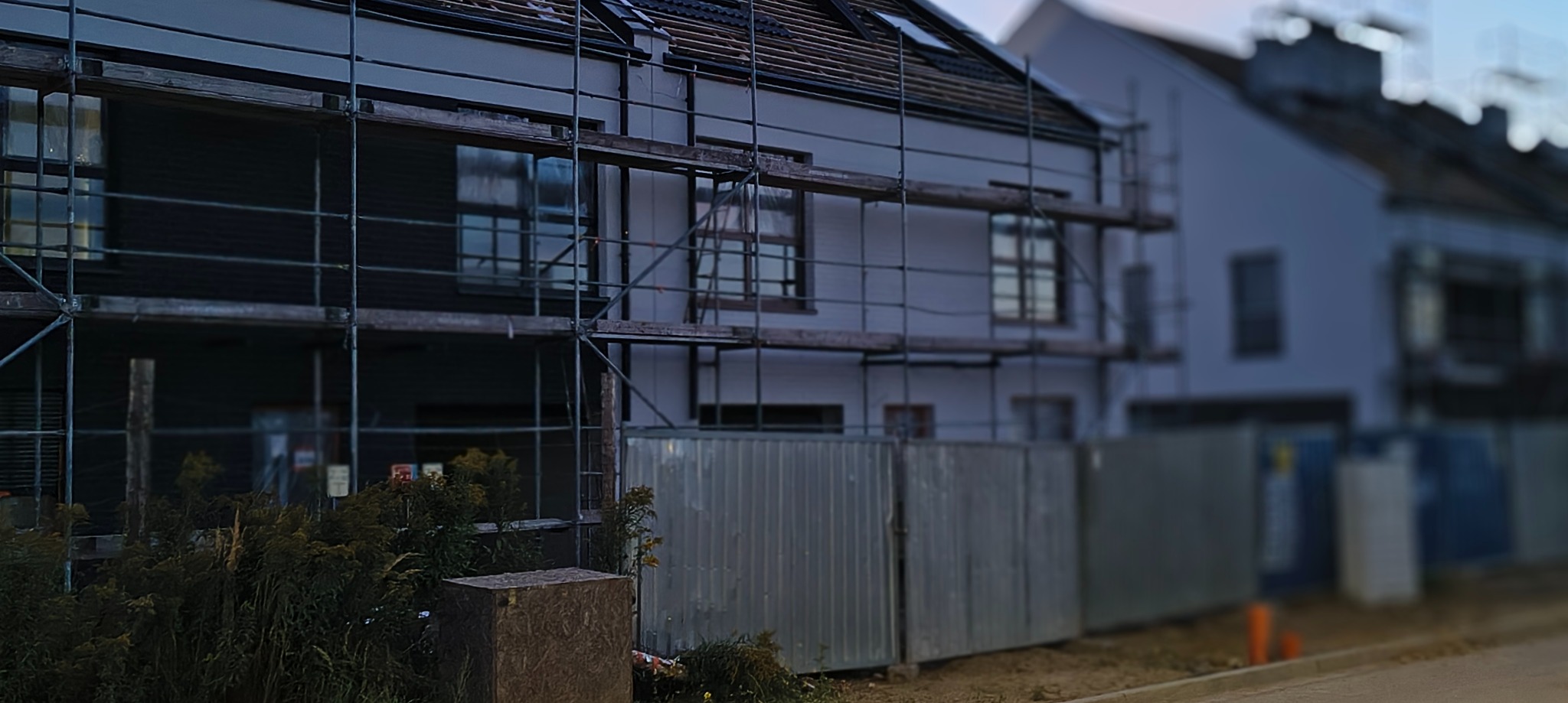 Dom w trakcie budowy/remontu, z rusztowaniem i ogrodzeniem. Widoczne okna i fragment dachu. Ujęcie z dołu, w tle inne budynki.