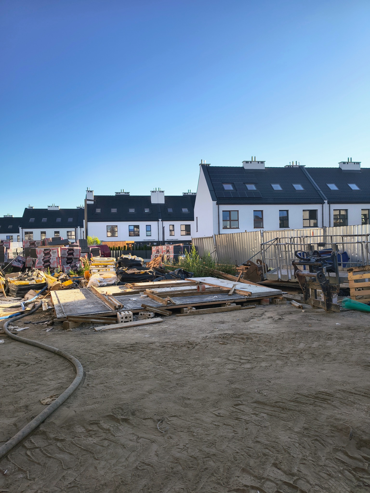 Plac budowy szeregowców z materiałami budowlanymi, widok z dołu na tle błękitnego nieba. Widoczne narzędzia i materiały przygotowane do prac wykończeniowych.