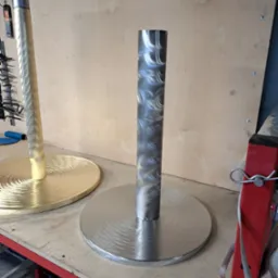 Dwie metalowe konstrukcje z okrągłymi podstawami, jedna srebrna, druga złota, z widocznymi śladami obróbki, na tle warsztatu.