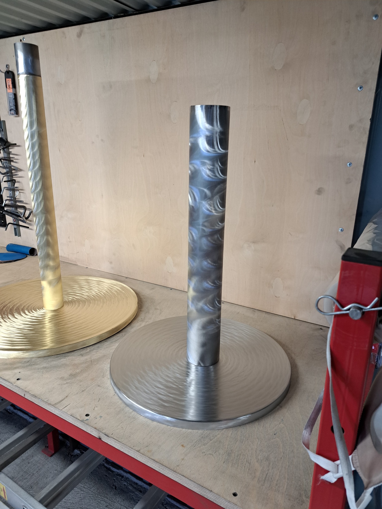 Dwie metalowe konstrukcje z okrągłymi podstawami, jedna srebrna, druga złota, z widocznymi śladami obróbki, na tle warsztatu.