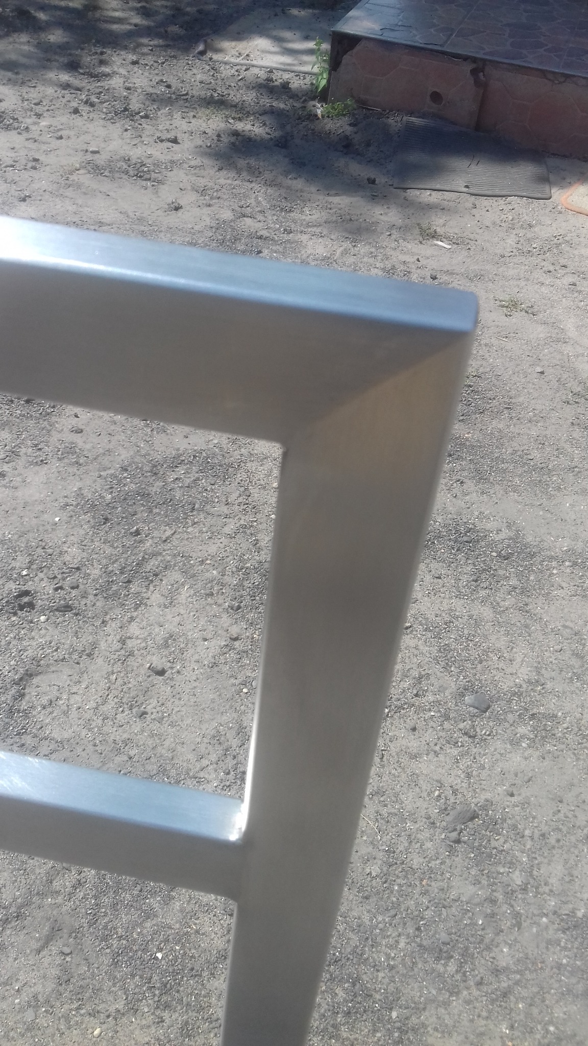 Fragment konstrukcji z nierdzewnej stali, spawana na zewnątrz. Widoczne łączenie profili, szlif i otoczenie z ziemią i fragmentem schodów.