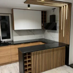 Nowoczesna kuchnia z drewnianymi lamelami i czarnym blatem. Białe szafki wiszące, oświetlenie punktowe. Wyspa z regałem na wino.