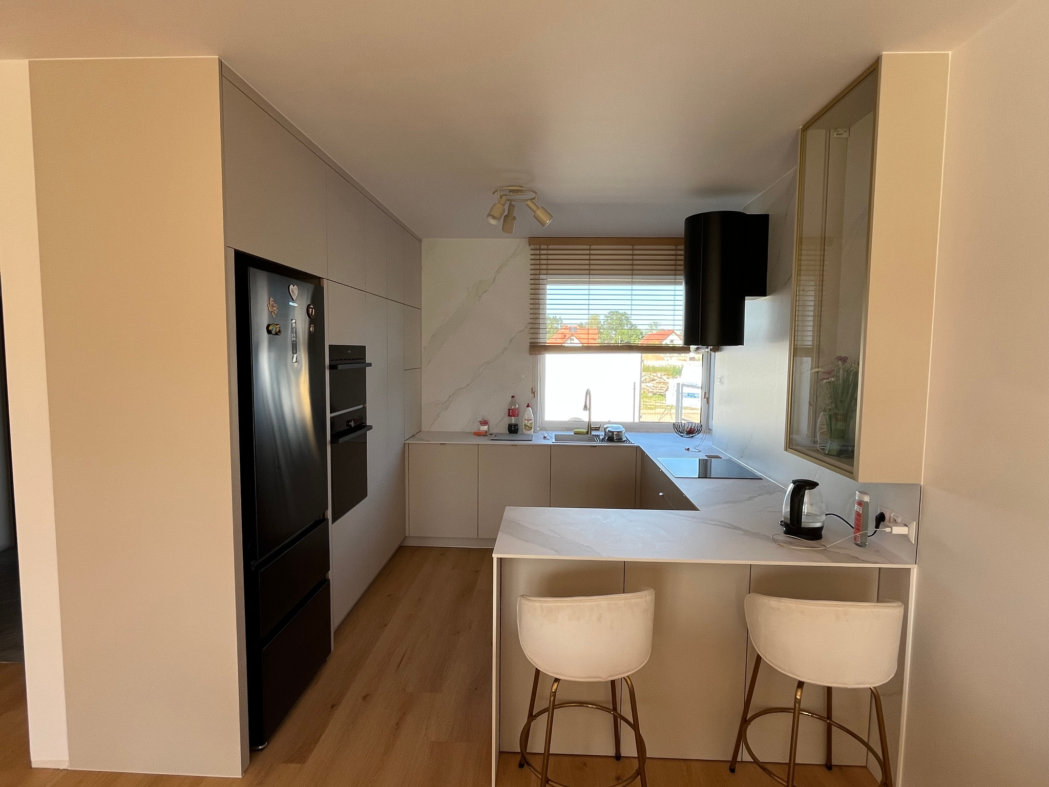 Jasna kuchnia w kształcie litery U z wyspą, beżowe fronty szafek, marmurowy blat i okładzina ściany, drewniane żaluzje w oknie, czarny sprzęt AGD, dwie białe hokery.