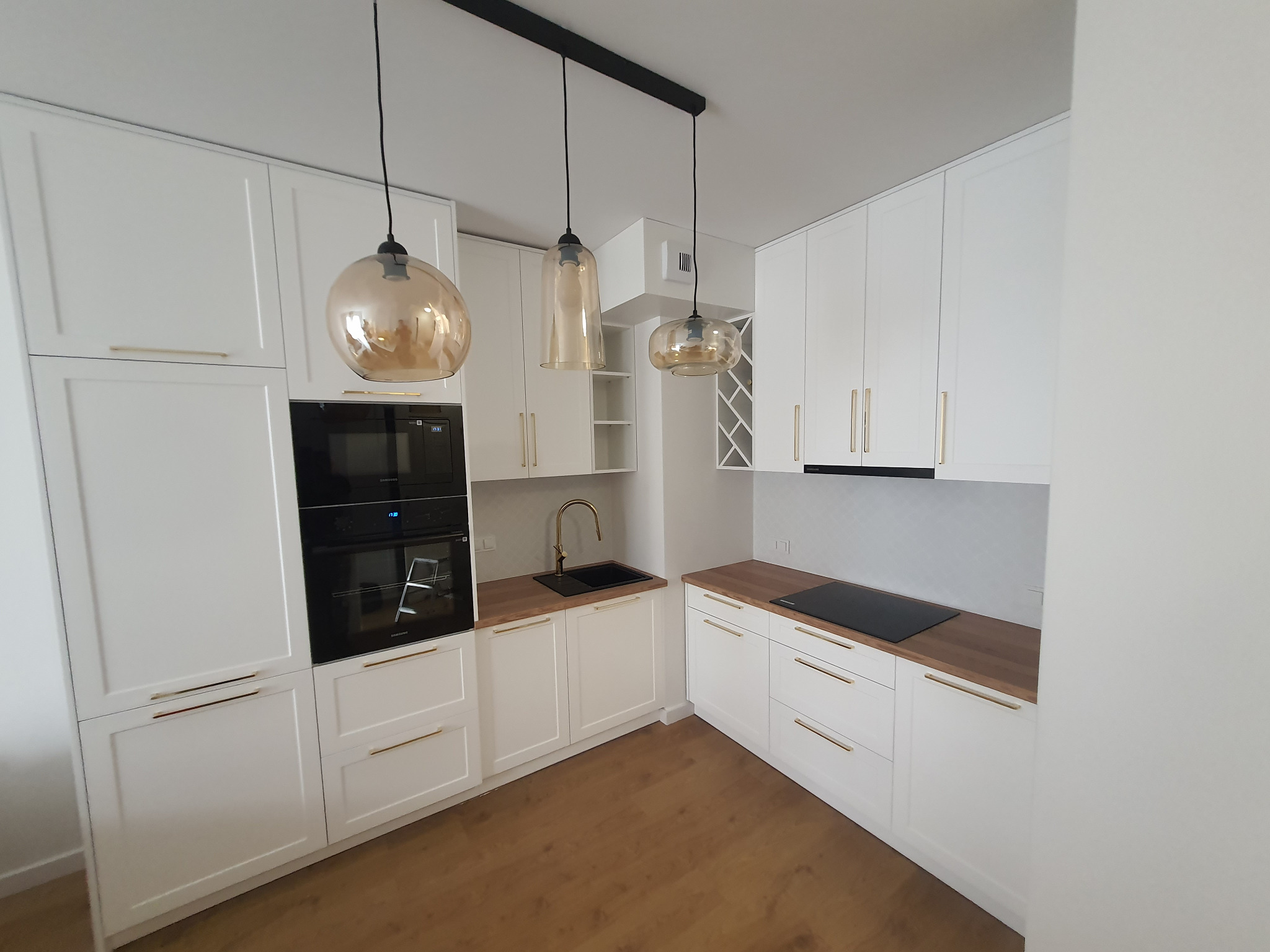 Biała kuchnia z drewnianym blatem i złotymi uchwytami, oświetlona trzema wiszącymi lampami o różnych kształtach, z piekarnikiem w zabudowie i czarnym zlewem.