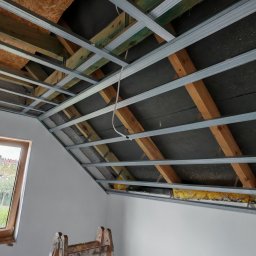Kamil Gałka - Sufit podwieszany z metalowym stelażem i izolacją, widok z dołu. Drewniana drabina oparta o ścianę, okno w tle. Prace wykończeniowe poddasza.