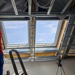 Kamil Gałka - Montaż stelaża pod płytę G-K na poddaszu z oknami dachowymi. Widoczny pracownik w czerwonej koszulce i drabina. W tle niebo przez okna.