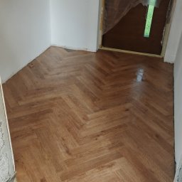 Kamil Gałka - Ułożona podłoga z paneli w jodełkę, widoczna przy białej ścianie i zabezpieczonych folią drzwiach, częściowo wykończona przy listwach.