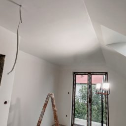 Kamil Gałka - Poddasze po malowaniu na biało, widoczna drabina, wiadro z farbą, lampa oświetleniowa i okno. Świeżo pomalowane ściany na biało.