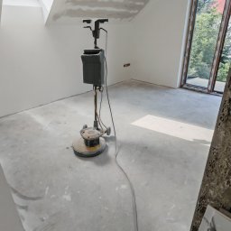 Kamil Gałka - Szlifowanie posadzki w nowym, jasnym pomieszczeniu z oknem. Na podłodze stoi maszyna do szlifowania. W tle widoczne ściany przygotowane do malowania.